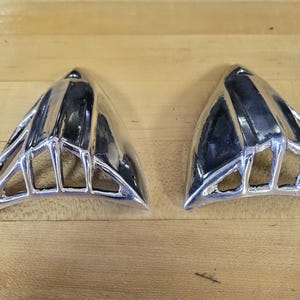 May include: Two silver-colored, triangular objects with a geometric design. The objects have a shiny, reflective surface and are placed on a light brown wooden surface. The design features a series of open, rectangular shapes.