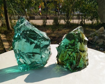 May include: Two pieces of green glass, one is a clear green and the other is a darker green with brown streaks. The glass pieces are sitting on a white surface.
