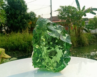 May include: A large, green, irregularly shaped piece of glass sits on a white surface. The glass is translucent and has a rough, textured surface.
