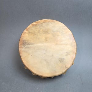 May include: A round, hand drum with a light tan, stretched skin surface. The drum's wooden frame is visible around the edges, and the drum is set against a gray background. The drum appears to be a traditional percussion instrument.