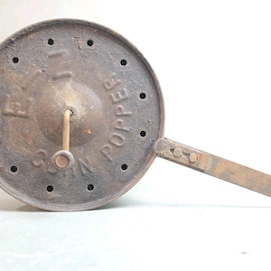 May include: Vintage metal corn popper with a round, perforated lid and a long handle. The lid is inscribed with the words "Corn Popper". The popper has a rustic, aged appearance.