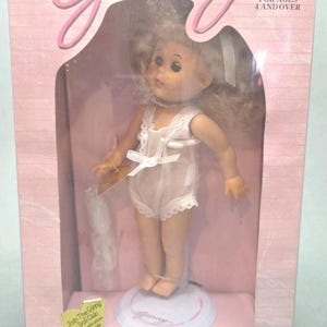 May include: A Ginny doll in a pink box. The doll is wearing a white romper with lace trim and a bow. The box has the words "Ginny" and "FOR AGES 4 AND OVER". The doll is on a white stand.