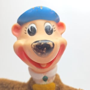 May include: A vintage cartoon bear figurine with a blue hat, a white collar, and a yellow shirt. The bear has a smiling expression with red lips and a black nose. The bear's fur is brown.