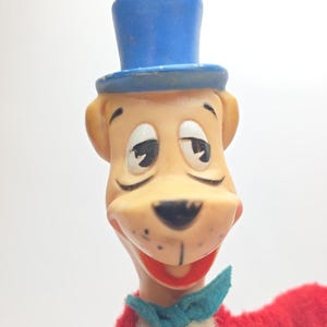 May include: A vintage cartoon dog puppet with a blue top hat and a teal bow tie. The dog has a tan face, black eyes, and a red mouth. The puppet's body is red and white.