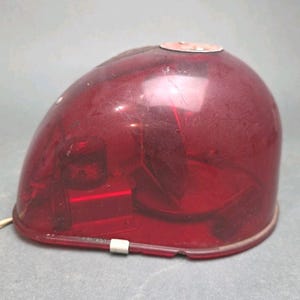 May include: A vintage, red, dome-shaped light fixture with a clear, plastic cover. The fixture has a white power cord and a metal base. The interior components are visible through the translucent cover. The light is likely from an emergency vehicle.