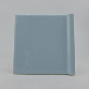 May include: A square, light blue ceramic tile with a glossy finish. The tile has a slightly curved edge on one side, adding a subtle design element. The tile is a solid color and appears to be new.