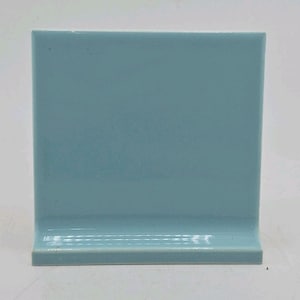 May include: A single, light blue ceramic tile with a glossy finish. The tile has a square shape with a raised edge along the bottom, creating a small shelf. The color is a muted, cool tone, suitable for various design applications.