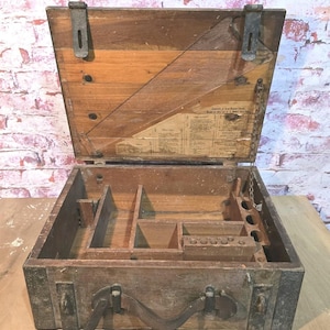 May include: Open wooden toolbox with a hinged lid and metal hardware. The interior features compartments and a handle for carrying. The box is made of aged wood with visible grain and a worn appearance, suggesting vintage use.