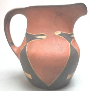 May include: A terracotta-colored pitcher with a dark handle and spout. The pitcher features a geometric design in black and gold, creating a striking contrast against the warm background. The design is symmetrical and adds a decorative element to the pitcher.