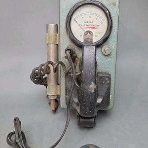 May include: Vintage EL-Tronics MR/HR radiation meter. The device has a green metal casing, a circular gauge, and a black handle. A black cord and a small black circular component are attached to the device.
