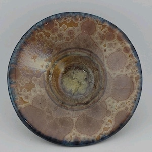 May include: A handmade ceramic bowl with a unique, textured surface. The bowl features a blend of colors, including brown, pink, and blue, creating a marbled effect. The rim has a dark blue glaze, and the interior has a lighter, speckled pattern.