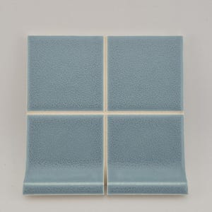 May include: Four square, light blue ceramic tiles with a textured surface. Two of the tiles have a curved edge. The tiles are arranged in a square formation, with a neutral background.
