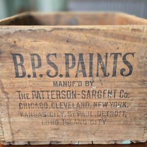 May include: A vintage wooden crate with the text "B.P.S. PAINTS" in black lettering. The crate also features the text "MANUF'D BY THE PATTERSON-SARGENT CO." and lists several cities. The crate is made of light brown wood.