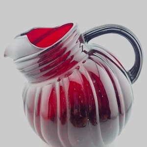 May include: A ruby red glass pitcher with a ribbed design. The pitcher has a curved handle and a spout. The interior of the pitcher is a bright red color. The pitcher is likely used for serving beverages.