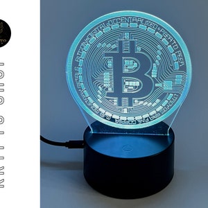 Puede incluir: Un logotipo de Bitcoin impreso en 3D azul y blanco sobre una base negra con un cable USB. El logotipo está iluminado con una luz azul. La base tiene un botón en la parte superior.
