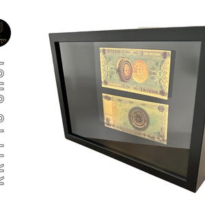 Peut inclure: Deux billets de Bitcoin encadrés avec des pièces d'or imprimées dessus. Les billets sont affichés dans un cadre noir avec un devant en verre.