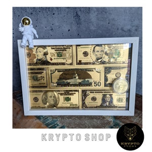 May include: A white frame containing a collage of gold-toned US dollar bills. The bills are arranged in a grid pattern and feature different denominations, including $1, $5, $10, $20, $50, and $100. A small astronaut figurine is perched on top of the frame.