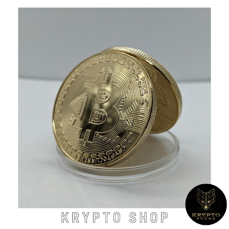 May include: A gold-colored Bitcoin coin with the Bitcoin symbol and text "PEER TO PEER * CRYPTOCURRENCY" on the front. The coin is on a clear plastic display stand.