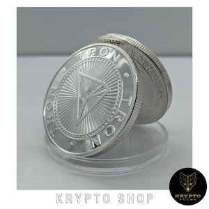 Könnte beinhalten: Nahaufnahme von zwei silbernen Münzen mit dem Wort "TRON" am Rand. Die Münzen haben ein dreieckiges Design in der Mitte mit radialen Linien. Die Münzen werden in einer transparenten Kunststoffhülle präsentiert. Der Text "KRYPTO SHOP" befindet sich unten.