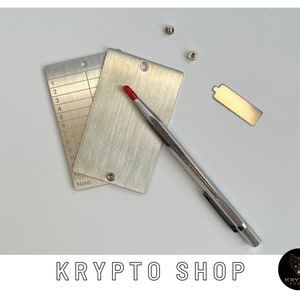 Può includere: Un set di tre strumenti in metallo argentato per testare l'autenticità delle monete. Gli strumenti includono un righello, una piccola piastra metallica e una penna argentata. Gli strumenti sono su una superficie bianca.