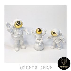 Könnte beinhalten: Drei weiße Astronautenfiguren mit goldenen Helmen. Der größte Astronaut winkt, der mittlere Astronaut sitzt auf einem weißen Mond und der kleinste Astronaut steht mit ausgestreckten Armen.