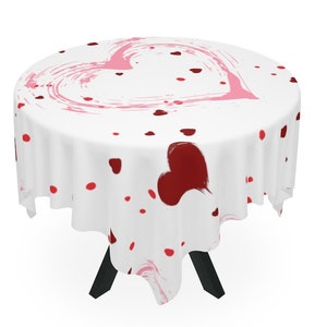 May include: White round tablecloth with a pink and red heart design. The tablecloth has a large pink heart in the center and smaller red hearts scattered around it.