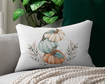 May include: A rectangular white throw pillow featuring a watercolor illustration of three stacked pumpkins in shades of cream, light blue, and orange, accented by green leaves and small pink berries arranged in a symmetrical botanical wreath design.