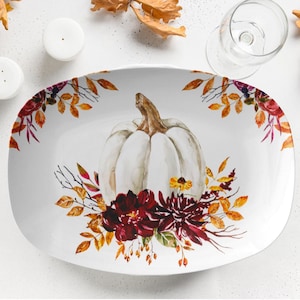 Puede incluir: Plato ovalado blanco con un diseño de acuarela de una calabaza blanca y un arreglo floral otoñal. El plato está decorado con hojas y flores burdeos, amarillas y naranjas, perfecto para reuniones de otoño.