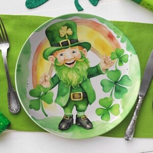May include: A white ceramic plate with a green shamrock design featuring a cartoon leprechaun with a green hat, beard, and clothing. The leprechaun is holding a pot of gold and smiling. The plate is surrounded by green shamrocks.