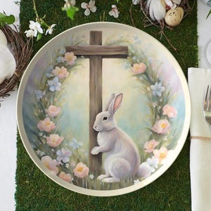 May include: A white ceramic plate with a painted design of a white rabbit standing next to a wooden cross. The rabbit is surrounded by a wreath of pink and blue flowers.