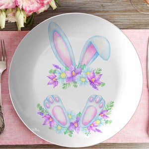 May include: White ceramic plate with a watercolor illustration of a bunny's ears and feet with purple and blue flowers.