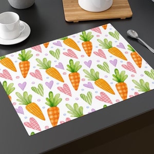May include: A white placemat with a repeating pattern of green leaves, orange carrots, and pink and purple hearts. The placemat is perfect for a spring or Easter dinner.