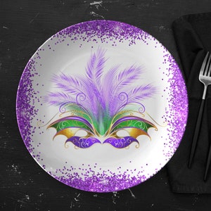 May include: A white ceramic plate with a purple glitter border. The center of the plate features a purple, green, and gold Mardi Gras mask with feathers.