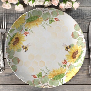 May include: A white ceramic dinner plate with a yellow and green floral design. The design features sunflowers, bees, and eucalyptus leaves. The plate has a honeycomb pattern in the center.