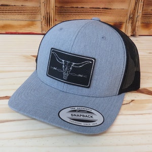 May include: A gray and black trucker hat with a black leather patch featuring a white skull with horns and barbed wire. The hat has a black mesh back and a silver oval sticker that reads "The Authentic Yupoong Snapback Since 1974".