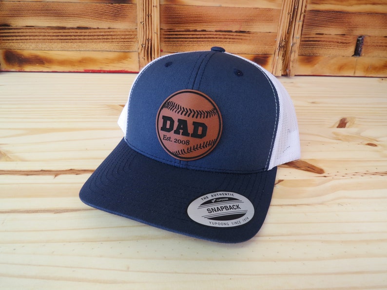 May include: A navy blue and white baseball cap with a brown leather patch that says "DAD Est. 2008" and a baseball graphic.