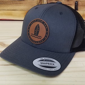 May include: A gray and black trucker hat with a brown leather patch that says "Surf and Sun California" with a surfboard graphic. The hat has a black mesh back and a snap closure.