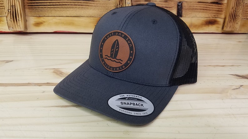 May include: A gray and black trucker hat with a brown leather patch that says "Surf and Sun California" with a surfboard graphic. The hat has a black mesh back and a snap closure.