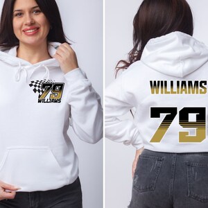 Op de afbeelding: Witte hoodie met een zwart en gouden nummer 79 en de naam "WILLIAMS" op de rug. De voorkant van de hoodie heeft een zwart-wit geblokt vlagontwerp met het nummer 79 en de naam "WILLIAMS" in goud.