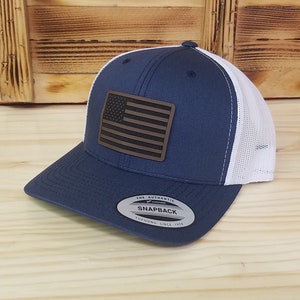 May include: A navy blue and white trucker hat with a brown leather American flag patch. The hat has a snap closure and a white mesh back.