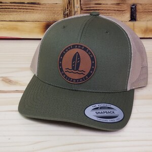 May include: A green and tan trucker hat with a brown leather patch that says "Surf and Sun California" with a surfboard graphic.