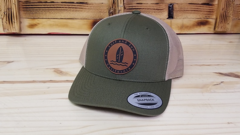 May include: A green and tan trucker hat with a brown leather patch that says "Surf and Sun California" with a surfboard graphic.