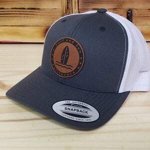 May include: A gray and white trucker hat with a brown leather patch that says "Surf and Sun California" with a surfboard graphic. The hat has a white mesh back and a black snap closure.