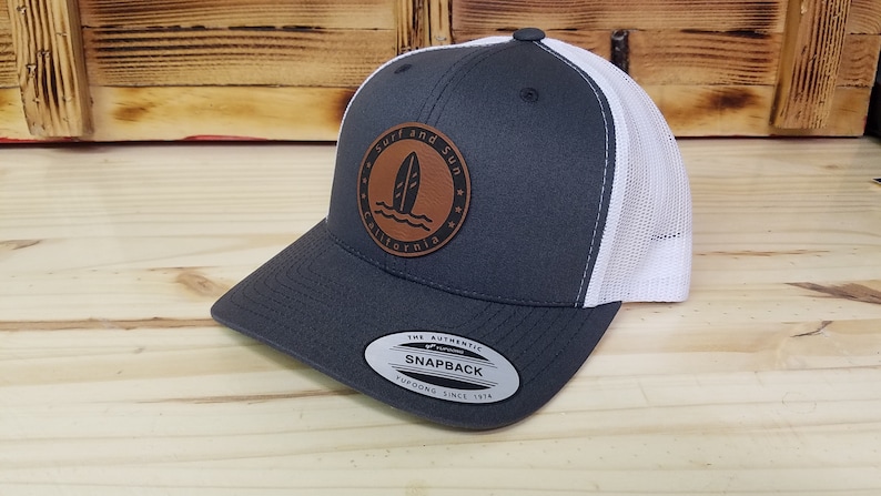 May include: A gray and white trucker hat with a brown leather patch that says "Surf and Sun California" with a surfboard graphic. The hat has a white mesh back and a black snap closure.