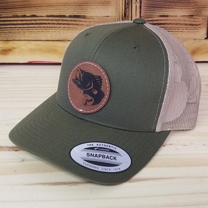 May include: A green and beige trucker hat with a brown leather patch featuring a black silhouette of a fish. The hat has a mesh back and a snap closure. The text "THE AUTHENTIC SNAPBACK" is printed on a white sticker on the front of the hat.