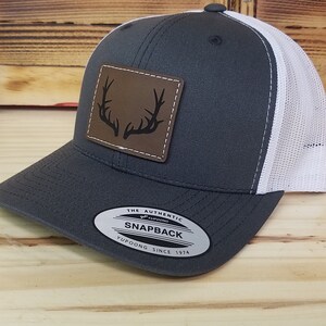 May include: A gray and white trucker hat with a brown leather patch featuring a black silhouette of antlers. The hat has a snap closure and a Yupoong logo on the back. The Authentic Snapback.