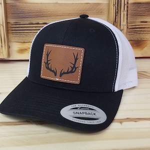 May include: Black and white trucker hat with a brown leather patch featuring a black silhouette of antlers. The hat has a white mesh back and a black front. The hat has a black snap closure and a white Yupoong logo sticker that says "The Authentic Snapback Yupoong Since 1974".