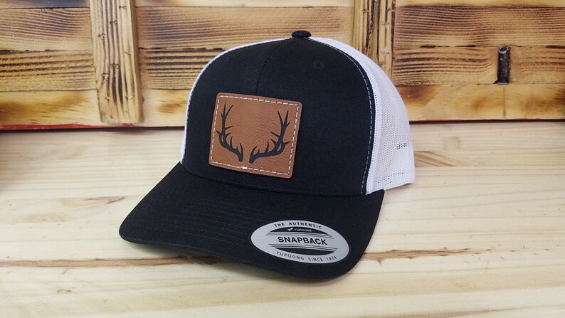 May include: Black and white trucker hat with a brown leather patch featuring a black silhouette of antlers. The hat has a white mesh back and a black front. The hat has a black snap closure and a white Yupoong logo sticker that says "The Authentic Snapback Yupoong Since 1974".