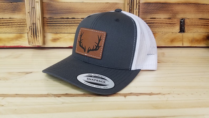 May include: Gray and white trucker hat with a brown leather patch featuring a black silhouette of antlers. The hat has a snap closure and a sticker that reads "The Authentic Snapback Yupoong Since 1974".