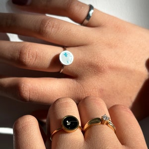 May include: Two gold rings with a small white flower design. One ring has a black background and the other has a white background.